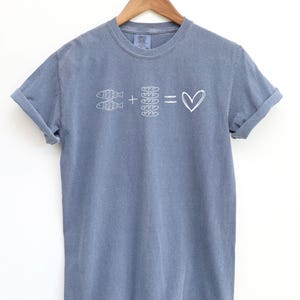 May include: A blue t-shirt with rolled-up sleeves. The shirt features a white graphic of two fish, bread, and a heart symbol. The design suggests a love for fish and bread.