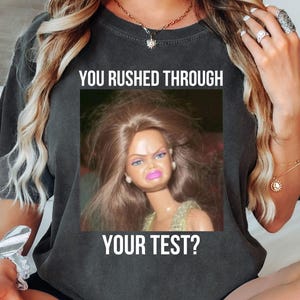 May include: Dark gray t-shirt with the text "YOU RUSHED THROUGH YOUR TEST?" above and below a photo of a doll with a displeased expression. The shirt is a crew neck.