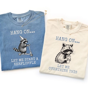 Könnte beinhalten: Zwei T-Shirts, eines in Blau und eines in Creme, mit Waschbär-Grafiken und Text. Das blaue Shirt sagt: "Hang On... Let Me Start A Kerfluffle." Das cremefarbene Shirt sagt: "Hang On... Let Me Overthink This."