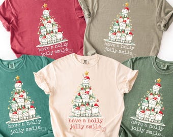 Könnte beinhalten: Fünf verschiedenfarbige T-Shirts mit einem Weihnachtsbaum-Design aus Zähnen mit Weihnachtsmützen. Der Text "have a holly jolly smile" ist unter dem Baum aufgedruckt. Die Shirts sind in Rot-, Grün- und Beigetönen gehalten.