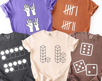 Puede incluir: Cinco camisetas de diferentes colores con diseños gráficos blancos. Los diseños incluyen gestos con las manos, marcas de conteo, un ábaco, cubos 3D y dados. Las camisetas son en tonos de morado, naranja, negro, crema y marrón.