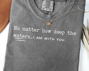 May include: A gray Comfort Colors t-shirt with the text "No matter how deep the waters...I AM WITH YOU." A gold-colored necklace with a circular pendant rests on the shirt. The shirt is folded and has a small tag.