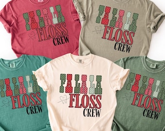 Könnte beinhalten: Fünf kurzärmelige T-Shirts in Rot-, Beige-, Türkis-, Creme- und Grüntönen. Jedes Shirt zeigt den Text "FA LA LA FLOSS CREW" mit Zahnseide-Picks und einer Zahngrafik. Das Design ist festlich und saisonal.