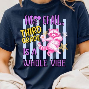 Puede incluir: Una camiseta azul marino de manga corta con un gr&aacute;fico de un mapache rosa sosteniendo una flor blanca. El texto dice: FIRST OF ALL... THIRD GRADE IS A WHOLE VIBE. El dise&ntilde;o incluye rayas verticales azul claro y estrellas amarillas y blancas.