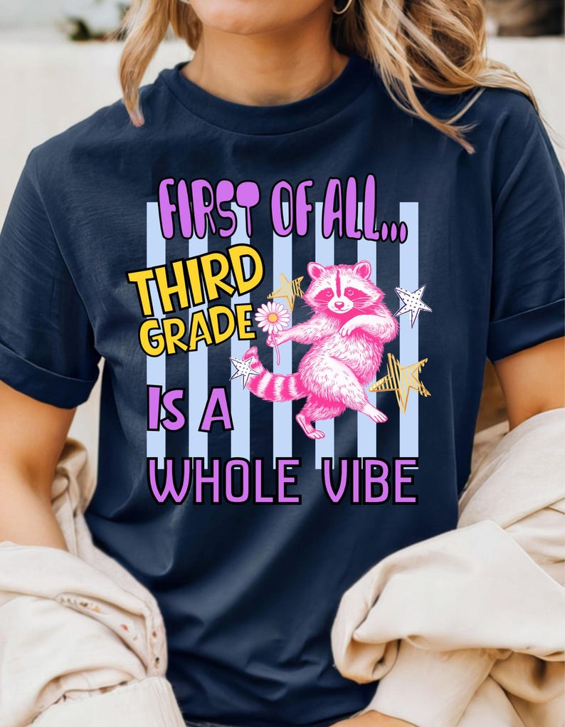 Puede incluir: Una camiseta azul marino de manga corta con un gr&aacute;fico de un mapache rosa sosteniendo una flor blanca. El texto dice: FIRST OF ALL... THIRD GRADE IS A WHOLE VIBE. El dise&ntilde;o incluye rayas verticales azul claro y estrellas amarillas y blancas.