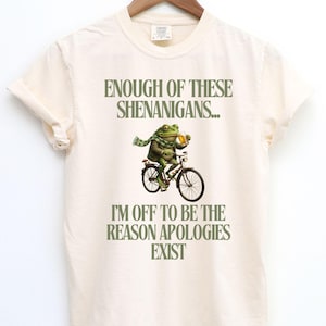 Könnte beinhalten: Cremefarbenes T-Shirt mit dem Text "ENOUGH OF THESE SHENANIGANS... I'M OFF TO BE THE REASON APOLOGIES EXIST." Ein Frosch mit Schal und Jacke fährt Fahrrad und hält ein Bier.