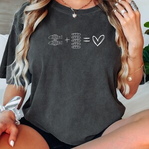 May include: Dark gray t-shirt with a white graphic. The graphic features two fish, plus four loaves of bread, equals a heart symbol. The shirt is a relaxed fit and has short sleeves. The model is wearing black shorts.