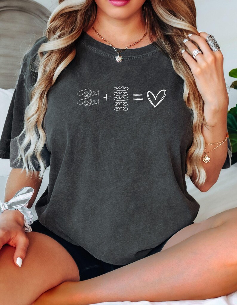 May include: Dark gray t-shirt with a white graphic. The graphic features two fish, plus four loaves of bread, equals a heart symbol. The shirt is a relaxed fit and has short sleeves. The model is wearing black shorts.