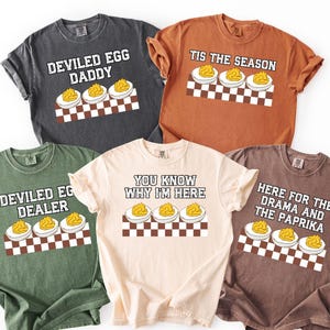Pode incluir: Cinco camisetas de cores diferentes com gráficos de ovos recheados e texto. As camisas são em tons de cinza, laranja, verde, bege e marrom. Cada camisa tem uma frase diferente, incluindo "Deviled Egg Daddy" e "Tis the Season."