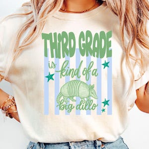 May include: Cream-colored t-shirt with the text "Third Grade is kind of a big dillo" in green and blue. The design includes an illustration of an armadillo and blue stars. The shirt is a casual style.