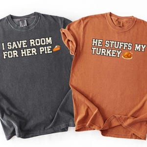 Könnte beinhalten: Ein anthrazitfarbenes T-Shirt mit dem weißen Text I SAVE ROOM FOR HER PIE und einer Tortengrafik sowie ein orangefarbenes T-Shirt mit dem weißen Text HE STUFFS MY TURKEY und einer Truthahngrafik, beide mit kurzen Ärmeln und Rundhalsausschnitt.