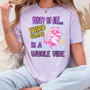 Puede incluir: Una camiseta de color morado claro con un gr&aacute;fico de un mapache rosa que sostiene una margarita, rodeado de estrellas amarillas y blancas. El texto dice "FIRST OF ALL... THIRD GRADE IS A WHOLE VIBE" en letras moradas, amarillas y negras.
