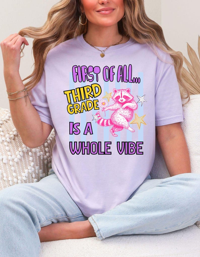Puede incluir: Una camiseta de color morado claro con un gr&aacute;fico de un mapache rosa que sostiene una margarita, rodeado de estrellas amarillas y blancas. El texto dice "FIRST OF ALL... THIRD GRADE IS A WHOLE VIBE" en letras moradas, amarillas y negras.