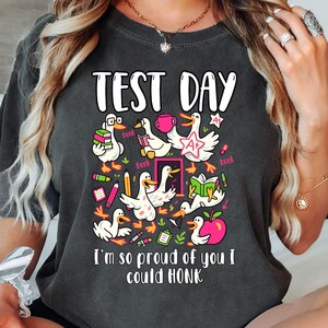 May include: Dark gray t-shirt with a colorful graphic featuring cartoon ducks with books, pencils, and an apple. The text "TEST DAY" is at the top, and "I'm so proud of you I could HONK" is at the bottom.