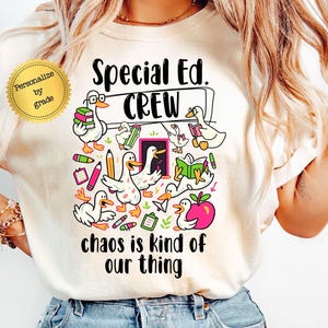 May include: Ivory-colored t-shirt with a graphic featuring cartoon ducks, books, and pencils. The text reads "Special Ed. CREW" and "chaos is kind of our thing." The design includes a pink apple and the option to personalize by grade.