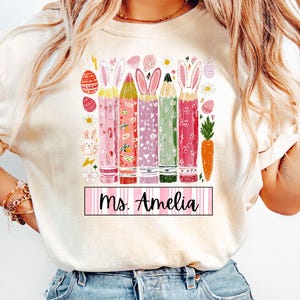 May include: Cream-colored t-shirt featuring a colorful graphic of Easter-themed pencils with bunny ears. The pencils are decorated with patterns and colors. The text "Ms. Amelia" is displayed on a pink and white striped banner.