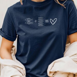 May include: Navy blue t-shirt with a white graphic. The graphic features two fish, plus four loaves of bread, equals a heart symbol. The shirt has short sleeves and a relaxed fit.