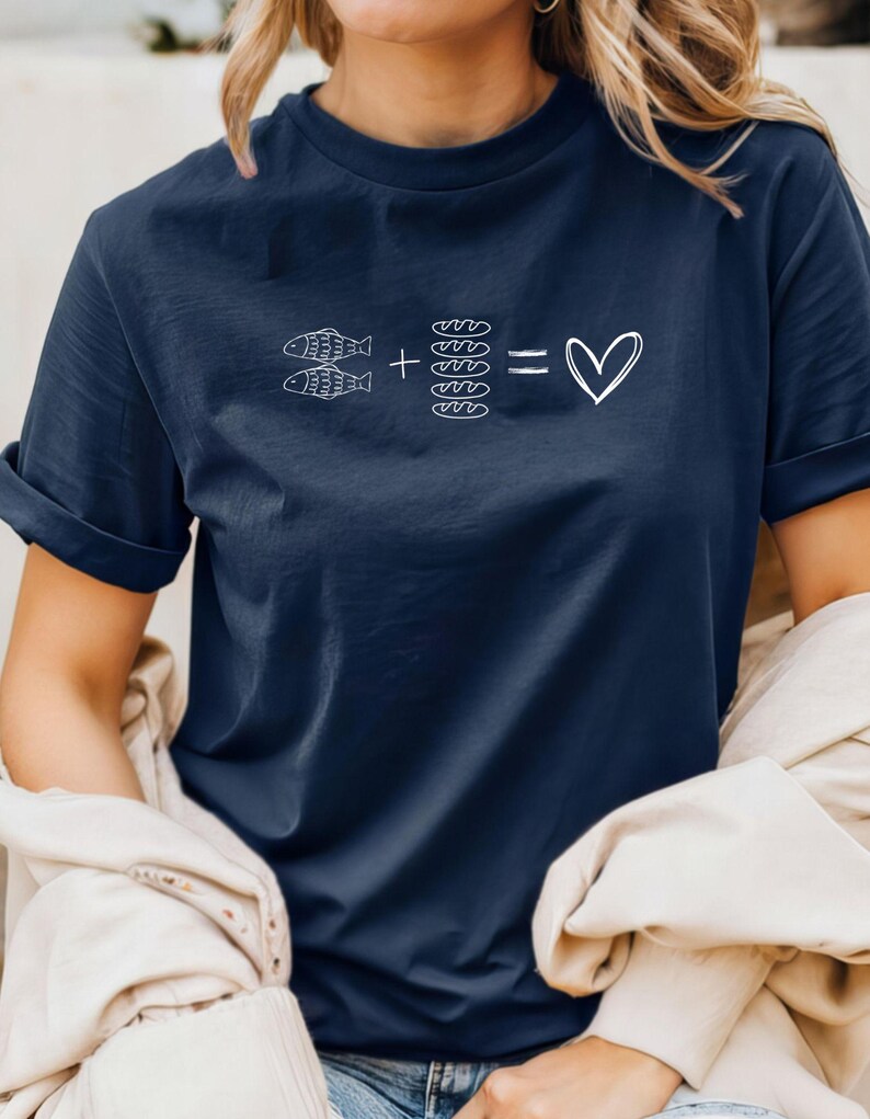 May include: Navy blue t-shirt with a white graphic. The graphic features two fish, plus four loaves of bread, equals a heart symbol. The shirt has short sleeves and a relaxed fit.