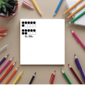 May include: White notepad with two ten-frame diagrams and the text "Ms. Miller" in black. The notepad is surrounded by colorful pencils, a small potted plant, and a pen on a light brown surface.