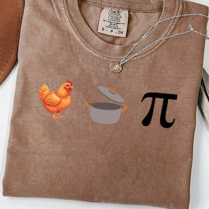 May include: Brown t-shirt featuring a chicken, a pot, and the pi symbol. The shirt has a crew neck and short sleeves. The brand label is visible on the upper left side of the shirt.