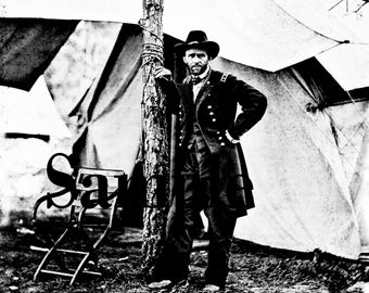 Pode incluir: Uma fotografia a preto e branco de um homem de uniforme militar de pé ao lado de uma árvore. Ele está usando um chapéu e tem barba. Ele está de pé em frente a uma tenda.