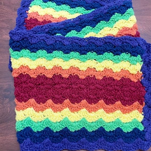 May include: A multi-colored crocheted blanket with a scalloped edge. The blanket is made in shades of blue, green, yellow, orange, and red.