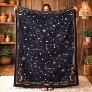 May include: A navy blue blanket featuring a celestial design with constellations and stars in gold. The blanket has a decorative border with crescent moons and starbursts. The soft blanket is held up in front of a wooden background.