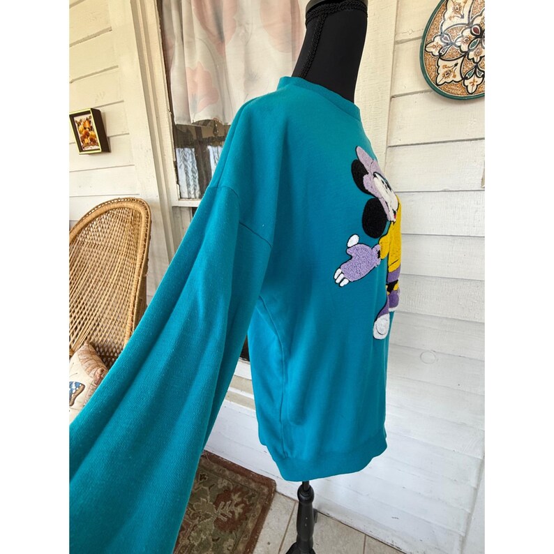 May include: Teal sweatshirt with a cartoon character design. The long-sleeved top has a crew neck and a graphic of a cartoon character in purple, yellow, and black. The sweatshirt is displayed on a mannequin.