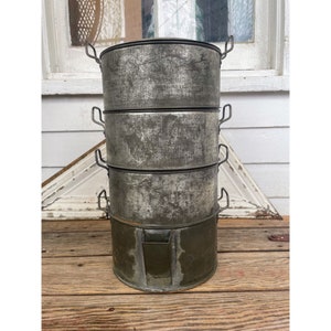 May include: A set of four vintage metal cake pans stacked on top of each other. The pans are silver with a distressed finish and have handles on the sides. The pans are sitting on a wooden surface.