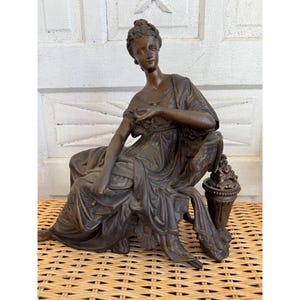 May include: Bronze sculpture of a seated woman in a flowing gown. The figure is posed on a detailed chair, with a small vase of flowers to the side. The sculpture has a classical aesthetic and is finished in a dark bronze color.