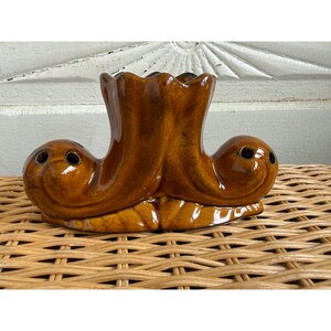 May include: A glossy brown ceramic flower frog featuring a central fluted opening and two rounded side sections, each with two small circular holes. The object has a smooth, glazed finish with darker brown shading in the recessed areas.