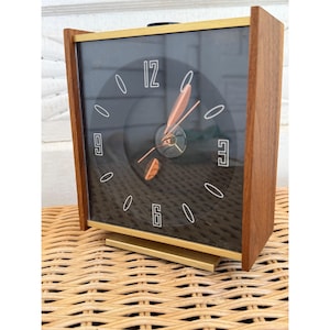 May include: A vintage rectangular clock with a black face, gold trim, and wooden sides. The clock face features white numerals and markers, with gold and copper-colored hands. The clock is set on a woven surface.