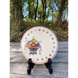 May include: A white ceramic plate with a floral design. The plate features a basket filled with colorful flowers and a border of small flowers and leaves.