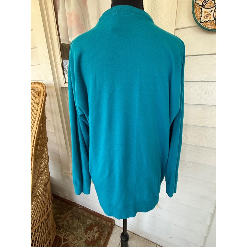 May include: A teal sweater with long sleeves, displayed on a mannequin. The fabric appears to be a soft knit. The sweater has a mock neck and a relaxed fit. The colour is a vibrant teal.