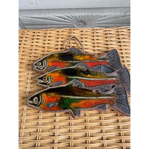May include: A decorative wall hanging featuring three stacked fish shapes crafted from enameled metal. Each fish displays a vibrant gradient of orange, yellow, green, and black colors with a glossy finish and a small metal loop for hanging.
