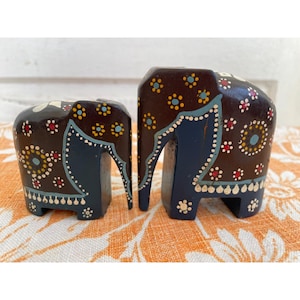 May include: Two hand-painted wooden elephant figurines. The larger elephant is brown with blue and white accents. The smaller elephant is brown with blue, white, and yellow accents.