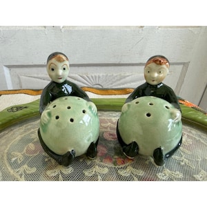 May include: A pair of ceramic salt and pepper shakers featuring two figures with dark green clothing, each holding a large, pale green, spherical container with multiple holes on top. The figures have painted facial features and brown hair.