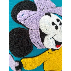 May include: Close-up of a teal sweatshirt featuring a textured Minnie Mouse design. The design includes a large purple bow, black ears, a white face, and a yellow top. The design is made with a textured, plush material.