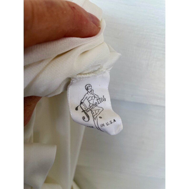 Op de afbeelding: Wit etiket met een zwart silhouet van een vrouw in een bikini en de tekst "Figurette" in een cursief schrift. Het etiket vermeldt ook "Made in U.S.A."