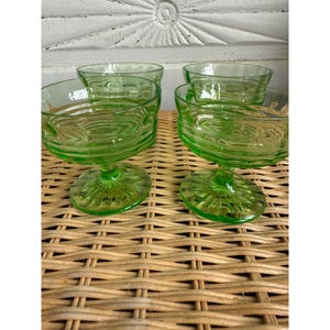 May include: Set of four green glass dessert or cocktail glasses. Each glass features a round bowl with a textured, concentric circle design and a pedestal base. The glasses are displayed on a woven surface.