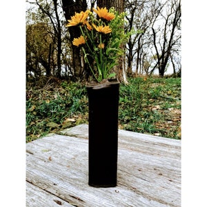 May include: A black, cylindrical vase made from repurposed materials, holding a bouquet of yellow flowers.