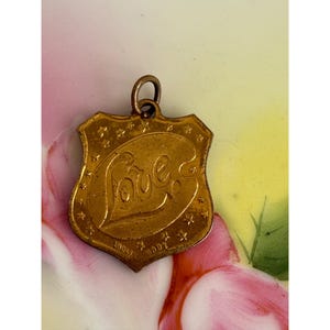 May include: A gold-colored pendant in the shape of a shield, with the word "Love" in cursive script. The pendant is adorned with small stars around the edges and the words "Made" and "With" at the bottom. The pendant has a small loop at the top for a chain.