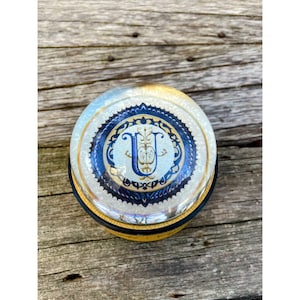 May include: A round, glass jar with a gold and blue lid. The lid features a decorative design with a gold letter "U" in the center.