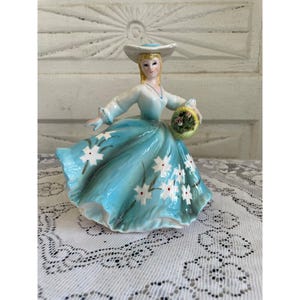 May include: A vintage porcelain figurine of a woman wearing a blue dress with white flowers and a white hat. She is holding a basket of flowers.
