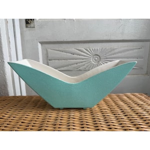 May include: A boat-shaped ceramic planter with a textured matte mint green exterior and a smooth white interior. The vessel features a sharp, angular silhouette with a low center dip and elevated pointed ends, supported by small feet at the base.