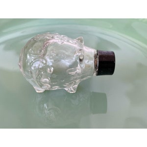 May include: A clear glass piggy bank with a black plastic stopper. The piggy bank has a detailed design of swirls and lines.
