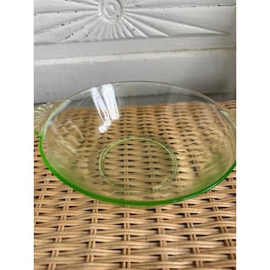 May include: A clear glass bowl with a green rim and handles. The bowl has a circular base and is set on a woven surface. The bowl is likely vintage or antique.