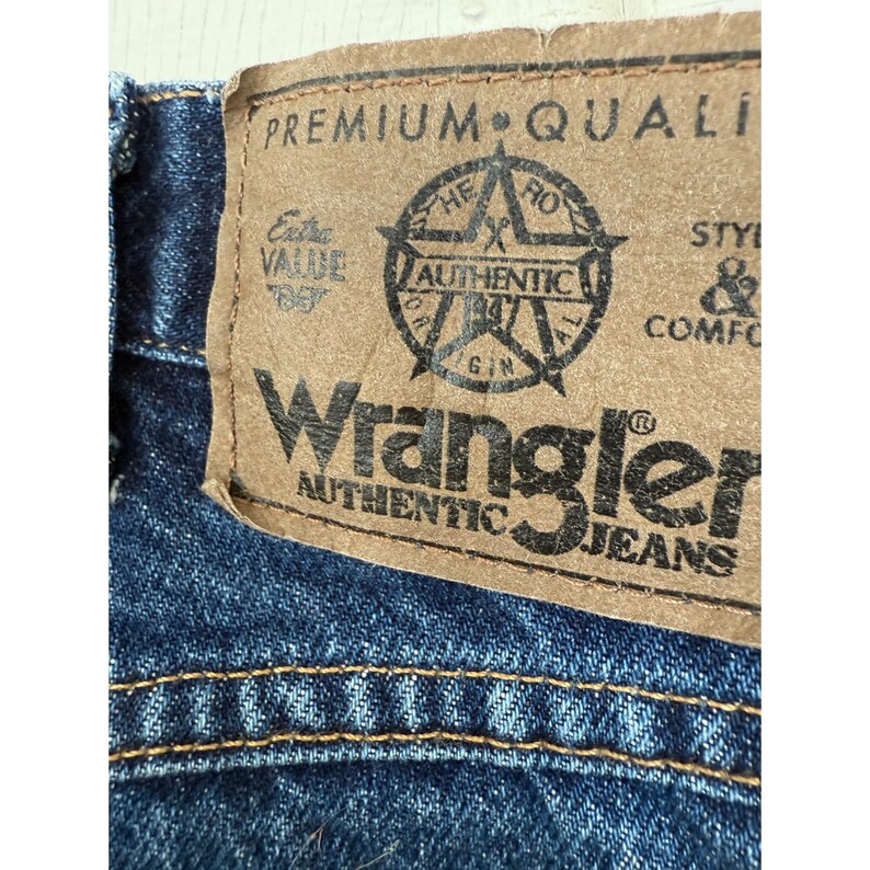 Puede incluir: Unos vaqueros de mezclilla azul oscuro con un parche de aspecto de cuero color canela en la cintura trasera. El parche muestra el texto Wrangler Authentic Jeans, Premium Quality y un logotipo de estrella con las palabras Hero, Authentic y Origin.