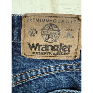 Puede incluir: Unos vaqueros de mezclilla azul oscuro con un parche de aspecto de cuero beige en la cintura trasera. El parche muestra el texto PREMIUM QUALITY, Wrangler AUTHENTIC JEANS, Extra VALUE, STYLE & COMFORT y un logotipo de estrella.