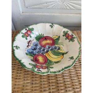 May include: A white ceramic fruit bowl with a scalloped edge and green trim. The bowl is decorated with a colorful illustration of various fruits, including apples, grapes, and bananas. The bowl is sitting on a woven surface.
